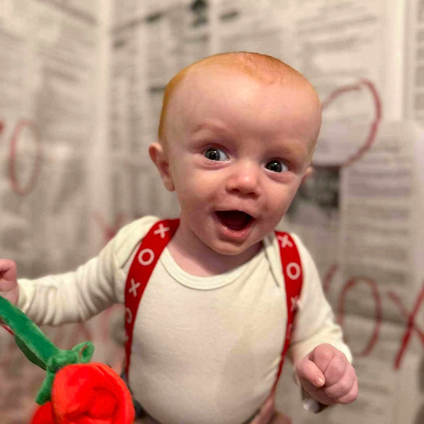 Jettson joined the competition — help win amazing prizes! baby, bokeh_background, close_up, cute, eyes, hand, happy, holding, indoor, infant, newspaper_wall, open_mouth, person, plush_rose, portrait, red_hair, smiling, surprised, suspenders, white_onesie