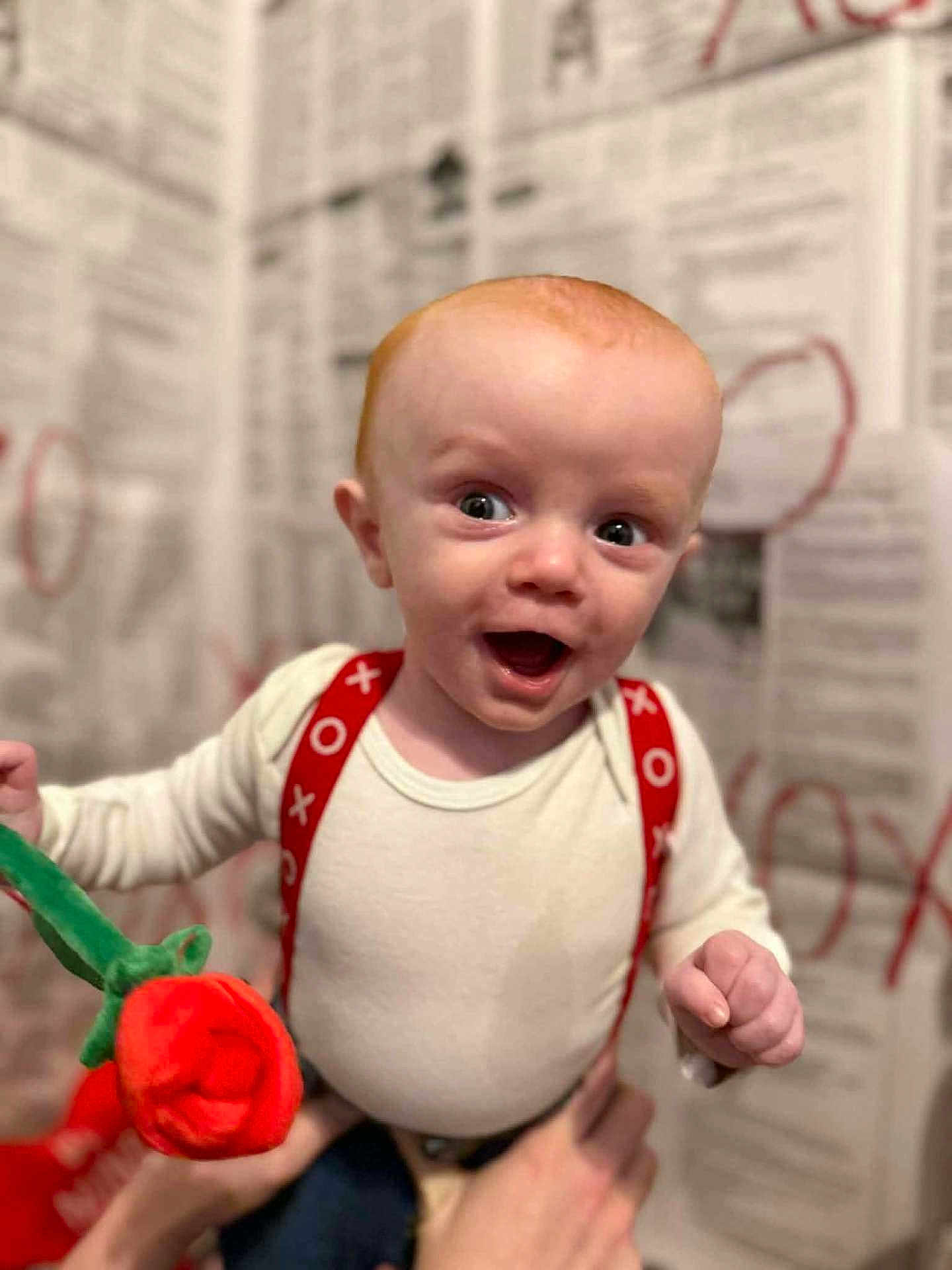 Jettson joined the competition — help win amazing prizes! baby, infant, smiling, surprised, red_hair, suspenders, white_onesie, plush_rose, hand, portrait, close_up, indoor, bokeh_background, newspaper_wall, eyes, open_mouth, cute, holding, person, happy
