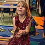 child, girl, dress, floral, chick, animal, classroom, rug, bee_pattern, backpack, temporary_tattoo, floor, furniture, indoor, happy, sitting, holding, curly_hair, playful, education