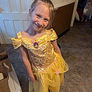 Ama is registered to the contest to win money with this photo: child, girl, princess_dress, yellow_dress, smile, indoor, tile_floor, playful, happy, dress_up, costume, footwear, flip_flops, long_hair, brown_hair, background, door, wall, paper_bags, room