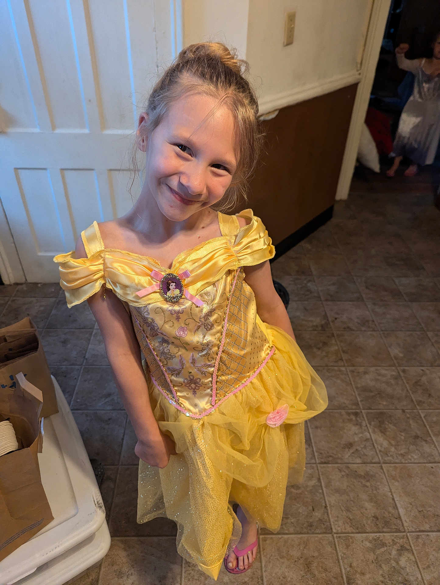 Ama is registered to the contest to win money with this photo: child, girl, princess_dress, yellow_dress, smile, indoor, tile_floor, playful, happy, dress_up, costume, footwear, flip_flops, long_hair, brown_hair, background, door, wall, paper_bags, room