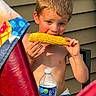 child, boy, corn, food, eating, shirtless, water_bottle, outdoor, summer, blue_eyes, blond_hair, table, chair, towel, sunlight, skin, hand, face, person, siding
