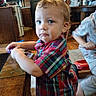 toddler, child, plaid_shirt, indoor, kitchen, table, wooden_table, carpet, playing_child, furniture, blue_eyes, curly_hair, person, young_child, home, casual_clothing, expression, standing, toy, background
