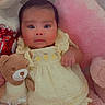 baby, teddy_bear, plush_toy, yellow_dress, pink_cushion, soft_toy, infant, cute, portrait, indoors, child, smiling, seated, furniture, cozy, adorable, young_child, comfortable, resting, home