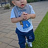 toddler, child, boy, outdoor, pavement, grass, bib, sneakers, short_hair, earrings, casual_clothing, curious_expression, daytime, building, fence, sky, clouds, walking, person, sidewalk