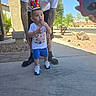 toddler, child, birthday_crown, toy, adult, tattoos, white_tshirt, blue_shorts, sneakers, outdoor, sunny, driveway, suburban, palm_trees, sidewalk, photography, person, hand, phone, casual