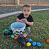 Gabriel is registered to the contest to win money with this photo: toddler, child, grass, bucket, plastic_eggs, outdoor, toy, black_shirt, shorts, shoes, earrings, wall, garden_bed, spring, easter, colorful, playful, curious, daylight, person