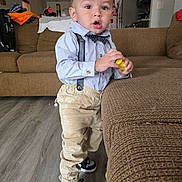 Gabriel is registered to the contest to win money with this photo: bow_tie, boy, casual, child, clothing, couch, curious, expression, floor, furniture, home, indoor, living_room, person, portrait, sneakers, standing, suspenders, toddler, toy