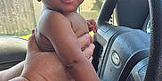 Amari is registered to the contest to win money with this photo: adult_hand, baby, blue_shirt, car_interior, child, curly_hair, daylight, diaper, eyes, face, fence, ford_logo, hand, house, jeans, person, seatbelt, skin, steering_wheel, window