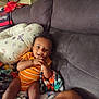 baby, child, couch, blanket, pillow, pacifier, beads, orange_onesie, mickey_mouse, smiling, indoor, happy, seated, curly_hair, furniture, cute, playful, comfort, home, person