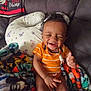 Amari joined the competition — help win amazing prizes! baby, blanket, child, couch, cozy, curly_hair, cute, happy, home, indoor, infant, orange_clothing, pacifier_clip, person, pillow, seated, smiling, striped_shirt, thumbs_up, toddler