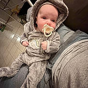 Emerson is registered to the contest to win money with this photo: baby, infant, pacifier, onesie, hooded_clothing, dinosaur_costume, person, lap, indoor, floor, tile_floor, furniture, blanket, couch, child, cute, soft_texture, home, casual, relaxing