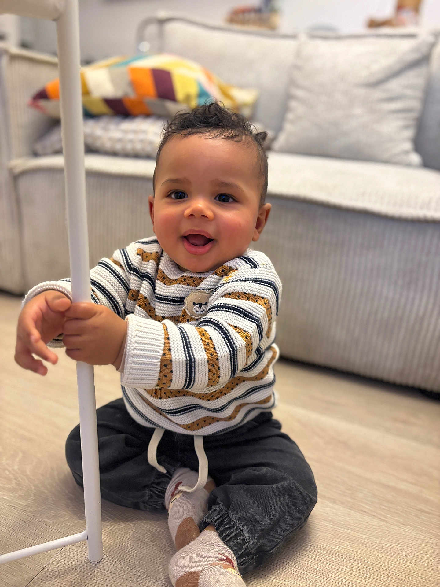 Isaiah participe au concours pour gagner de l'argent avec cette photo : bear_pattern, child, colorful_pillow, couch, cozy, curly_hair, cushions, cute, dark_pants, happy, home, indoor, portrait, sitting, smiling, socks, striped_sweater, toddler, white_pole, wooden_floor