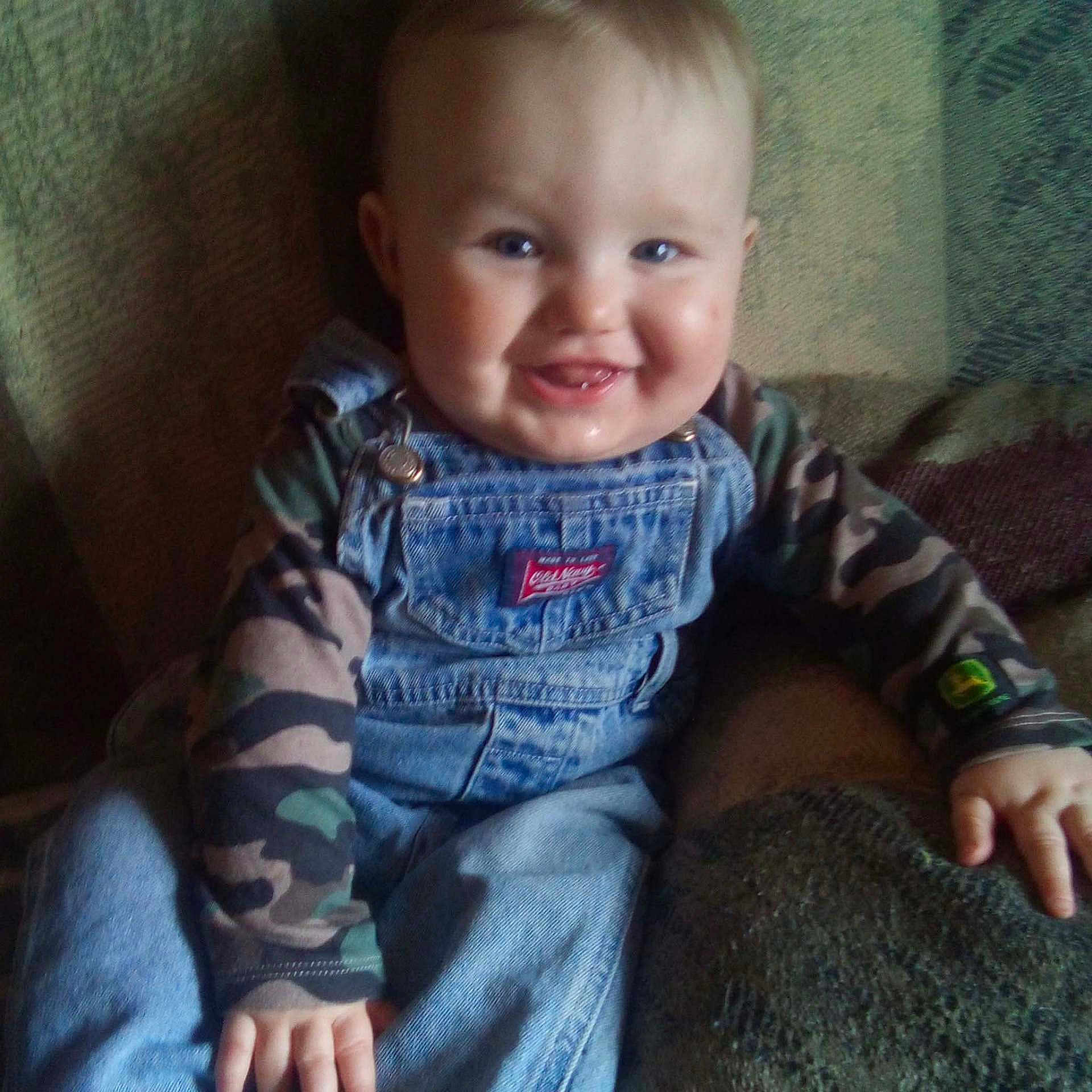 James joined the competition — help win amazing prizes! baby, child, smiling, denim_overalls, camo_clothing, sitting, couch, happy, face, person, indoor, cute, cheeks, teeth, hand, blue_eyes, clothing, textile, portrait, infant