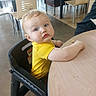 baby, blue_eyes, casual, child, curious, dining_area, expression, face, floor, furniture, high_chair, indoor, light, looking, person, restaurant, seated, table, wooden_table, yellow_shirt