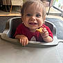 toddler, child, high_chair, food, snack, red_shirt, indoor, smiling, happy, hair, face, hand, table, seat, window, light, floor, restaurant, person, cute