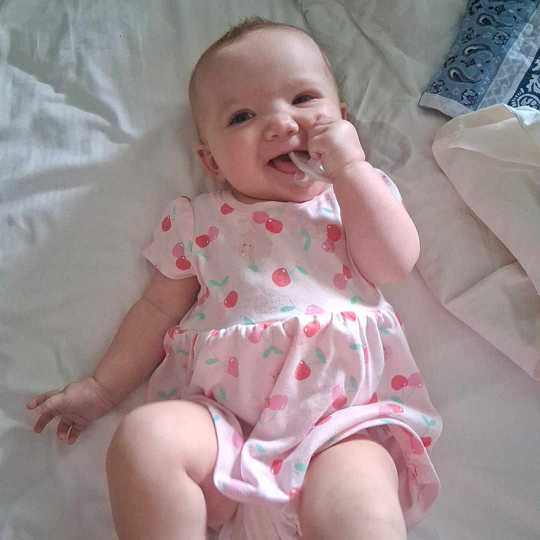 Lirosea is registered to the contest to win money with this photo: baby, bed, bedding, cherry_pattern, child, cute, hand, happy, indoor, infant, legs, lying_down, person, pink_dress, skin, sleepwear, smiling, soft, teething_toy, young_child