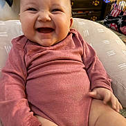 Riley is registered to the contest to win money with this photo: baby, child, infant, smiling, laughing, happy, portrait, close_up, face, eyes, open_mouth, pink_outfit, onesie, seated, cushion, indoor, home, dresser, clothing_basket, cozy