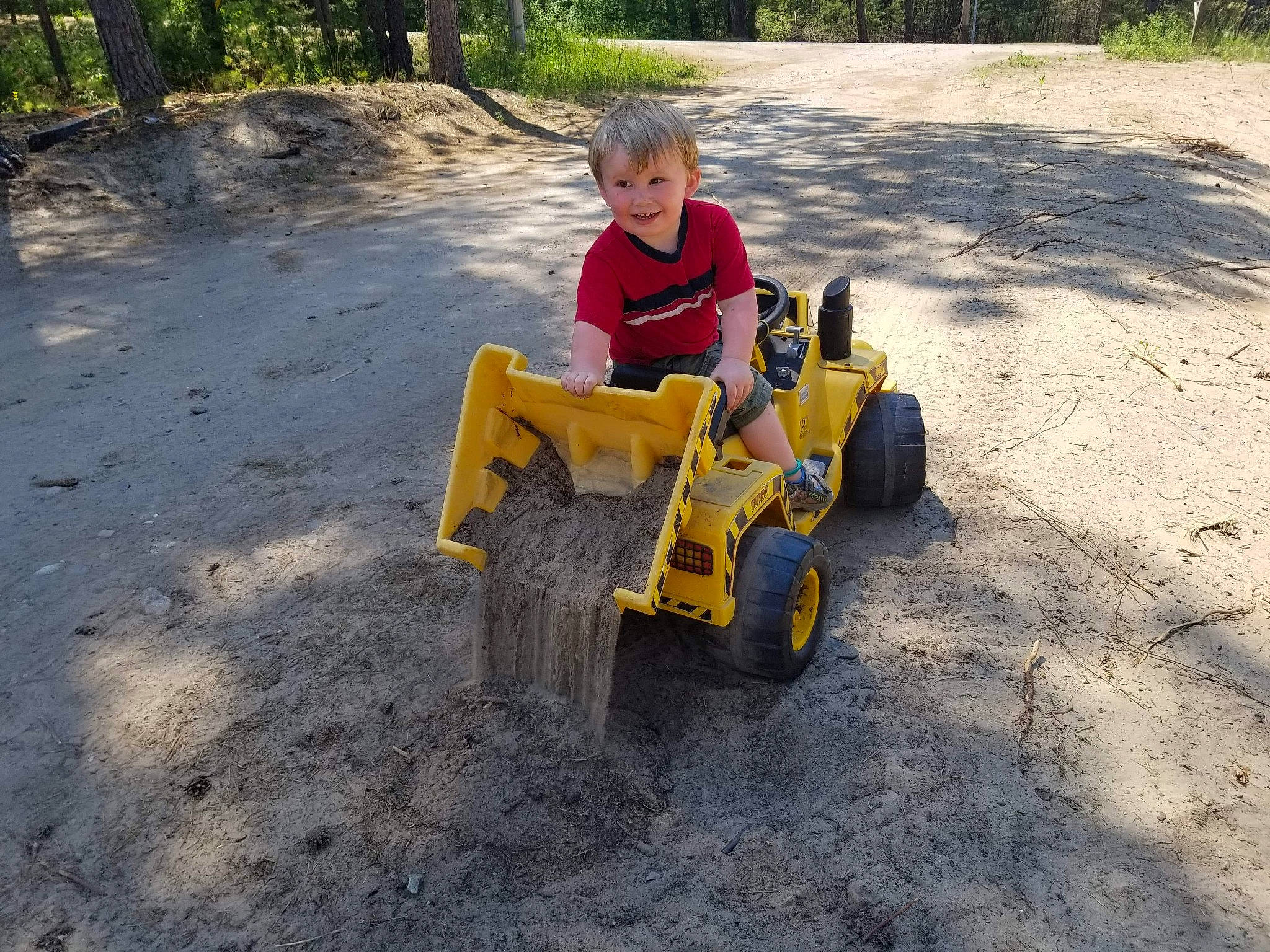 Louis is registered to the contest to win money with this photo: asphalt, child, construction_equipment, joy, person, play, riding_toy, soil, toddler, tree, vehicle, wheel