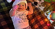 Giovanni is registered to the contest to win money with this photo: baby, blanket, child, christmas_lights, christmas_tree, cozy, cute, decorations, festive, floor, happy, holiday, indoors, infant, pajamas, person, plush_toy, santa_hat, smiling, toy