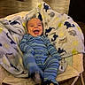 baby, infant, smiling, blue_pajamas, blanket, animal_pattern, baby_rocker, indoor, floor, wooden_floor, happy, cozy, child, person, clothing, furniture, home, cute, sitting, playful