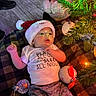 Giovanni is registered to the contest to win money with this photo: baby, santa_hat, christmas_tree, ornament, blanket, holiday, infant, floor, lights, plush_toy, clothing, cute, festive, decorations, indoors, person, toddler, patterned_pants, smiling, celebration