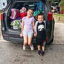 children, kids, car_trunk, backpack, bags, smiling, outdoor, casual_clothes, playful, sibling, holding_hands, daytime, concrete, residential_area, greenery, summer_clothes, footwear, happy, family, transportation