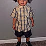 toddler, child, curly_hair, plaid_shirt, shorts, sneakers, indoor, carpet, wall, standing, portrait, person, cute, young, smiling, fashion, casual, feet, hands, expression
