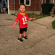 Xander is registered to the contest to win money with this photo: toddler, child, red_jersey, sportswear, shorts, sneakers, concrete, driveway, brick_house, window, greenery, grass, sidewalk, curious_expression, outdoor, daylight, person, casual_clothing, young_child, standing