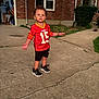 toddler, child, red_jersey, sportswear, shorts, sneakers, concrete, driveway, brick_house, window, greenery, grass, sidewalk, curious_expression, outdoor, daylight, person, casual_clothing, young_child, standing