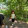 child, toddler, forest, trail, trees, greenery, outdoor, nature, sneakers, tank_top, person, water_bottle, path, sunlight, shadows, curly_hair, casual_clothing, daylight, walking, young_child