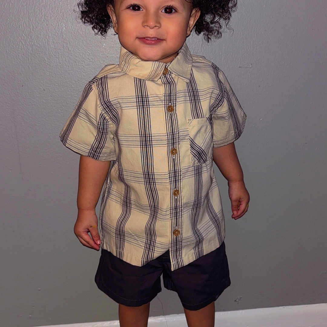 Xander is registered to the contest to win money with this photo: black_shorts, carpet, casual_clothing, child, curly_hair, cute, fashion, front_view, full_body, gray_wall, happy, indoor, person, plaid_shirt, portrait, smiling, sneakers, standing, toddler, young_child