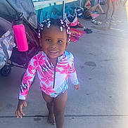 Kiara is registered to the contest to win money with this photo: child, girl, swimsuit, beads, hair, smile, barefoot, stroller, water_bottle, poolside, concrete, people, reflection, bag, towel, summer, outdoor, sunlight, casual, playful