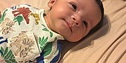 Makari is registered to the contest to win money with this photo: baby, infant, dinosaur_clothing, smiling, lying_down, bed, hand, soft_surface, cute, portrait, child, indoors, cozy, colorful, sleepwear, young_child, person, fabric, relaxed, skin