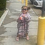 child, girl, sunglasses, sidewalk, plaid_shirt, sandals, yellow_bollard, parking_lot, cars, smile, playful, pigtails, outdoor, concrete, street, wet_surface, person, clothing, accessory, urban