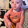 baby, infant, child, toy, colorful_toy, orange_clothing, cushion, blanket, patterned_blanket, plaid_pattern, cow_print, indoor, seated, curious_expression, blue_eyes, short_hair, skin, hand, foot, soft_fabric