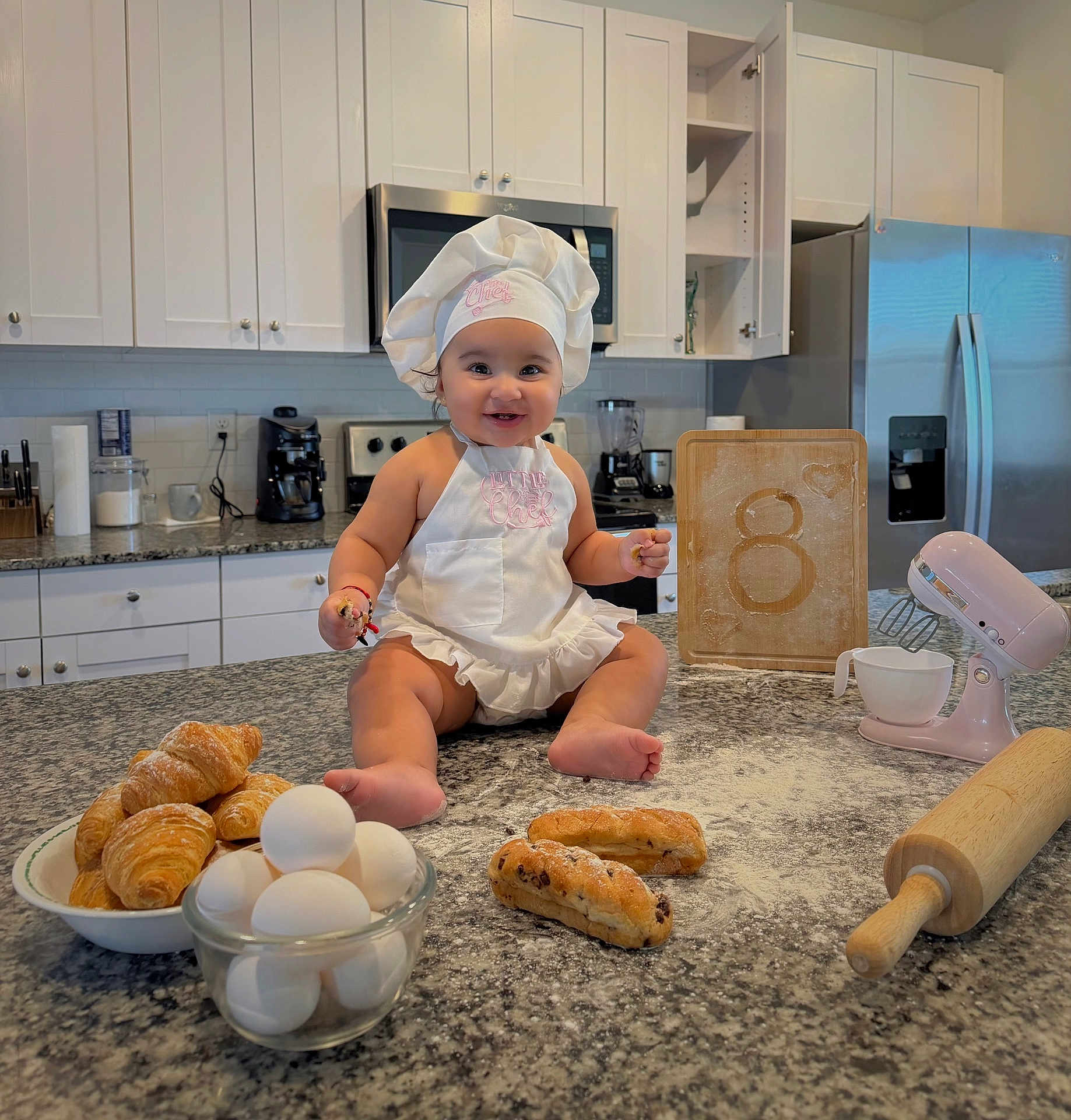 Madisson joined the competition — help win amazing prizes! baby, chef_hat, apron, kitchen, countertop, eggs, croissants, bread, rolling_pin, mixer, cutting_board, flour, smile, happy, cooking, baking, infant, home, food, portrait