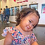 child, girl, curly_hair, smile, floral_dress, frozen_yogurt_shop, indoor, cute, person, young_child, seated, playful, happy, portrait, colorful, casual_clothing, head_tilt, people, background, food_shop