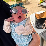 Léonie participe au concours pour gagner de l'argent avec cette photo : baby, sunglasses, headband, hand, adult_hand, outdoor, clothing, person, child, table, bag, stone_wall, greenery, flower_pattern, knitted_sweater, surprised_expression, holding, summer, casual, portrait