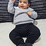 Aariz is registered to the contest to win money with this photo: baby, smiling, child, infant, grey_sweater, black_pants, socks, lying_down, pillow, soft_texture, indoors, cute, happy, person, face, head, raised_hand, cozy, blanket, portrait