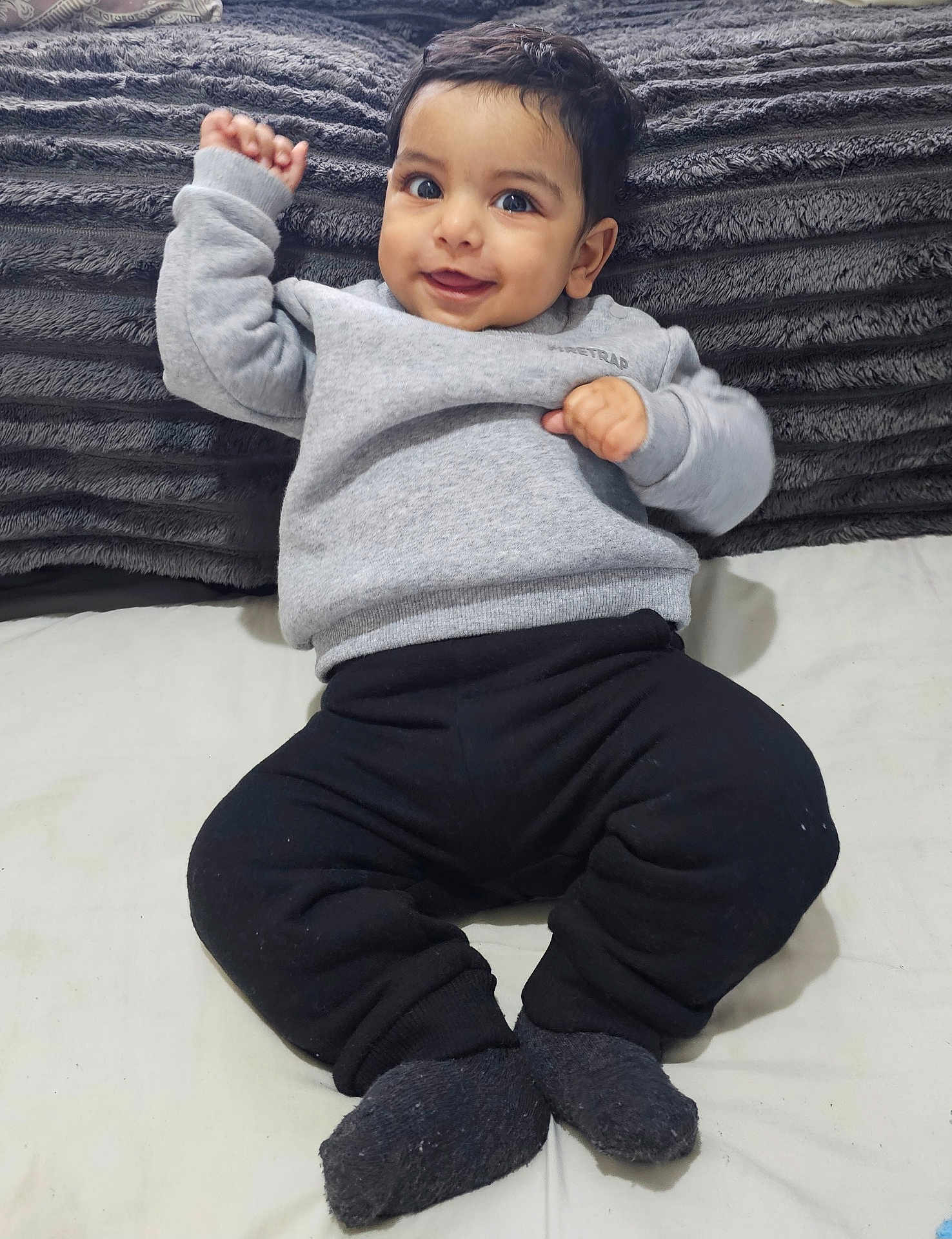 Aariz is registered to the contest to win money with this photo: baby, smiling, child, infant, grey_sweater, black_pants, socks, lying_down, pillow, soft_texture, indoors, cute, happy, person, face, head, raised_hand, cozy, blanket, portrait