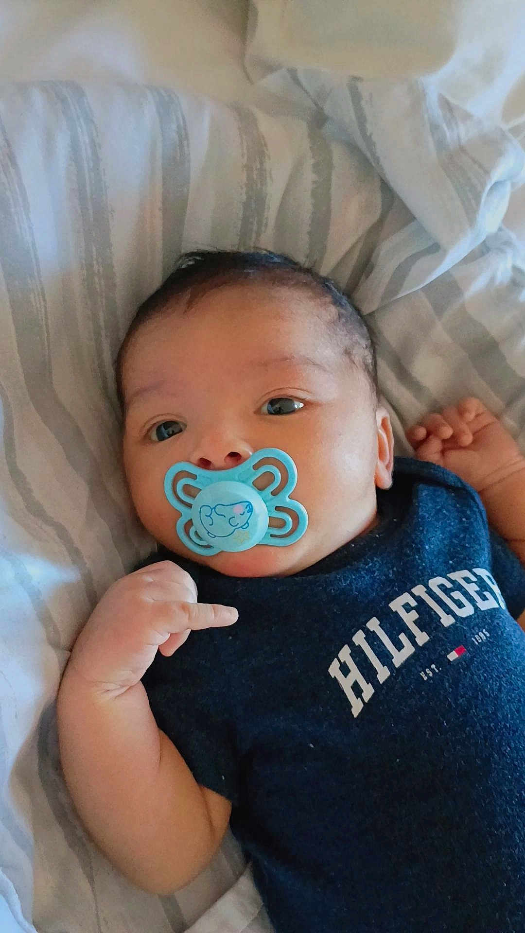 Josué Leonardo is registered to the contest to win money with this photo: baby, pacifier, infant, blue_pacifier, clothing, bed, striped_bed_sheet, hand, face, eyes, cute, lying_down, navy_shirt, hilfiger, child, person, skin, young_child, indoor, portrait