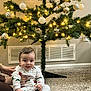 baby, child, pajamas, carpet, christmas_tree, ornaments, lights, indoor, holiday, festive, smiling, person, hand, floor, decorations, home, cozy, cheerful, sitting, warm_lighting
