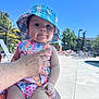 baby, child, swimsuit, hat, plaid, floral, poolside, adult_hand, outdoor, sunny, blue_sky, fence, lounge_chair, person, summer, vacation, cute, infant, daytime, portrait