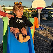 Arrow Saige is registered to the contest to win money with this photo: toddler, child, slide, playground, outdoor, basketball_hoop, hand, black_clothing, orange_bows, curly_hair, sunlight, toys, garden, plants, wood_chips, boots, person, daytime, expression, fence