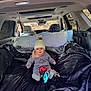 baby, child, car_interior, blanket, pillow, toy, hat, winter_clothing, pom_pom, sitting, cozy, soft_toy, car_seat, window, sunroof, seatbelt, gray_clothing, indoor, portrait, smiling