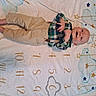 baby, barefoot, blanket, child, closeup, face, happy, indoor, infant, khaki_pants, lying_down, numbered_blanket, nursery, photograph, plaid_shirt, playful, portrait, smiling, star_pattern, suspenders