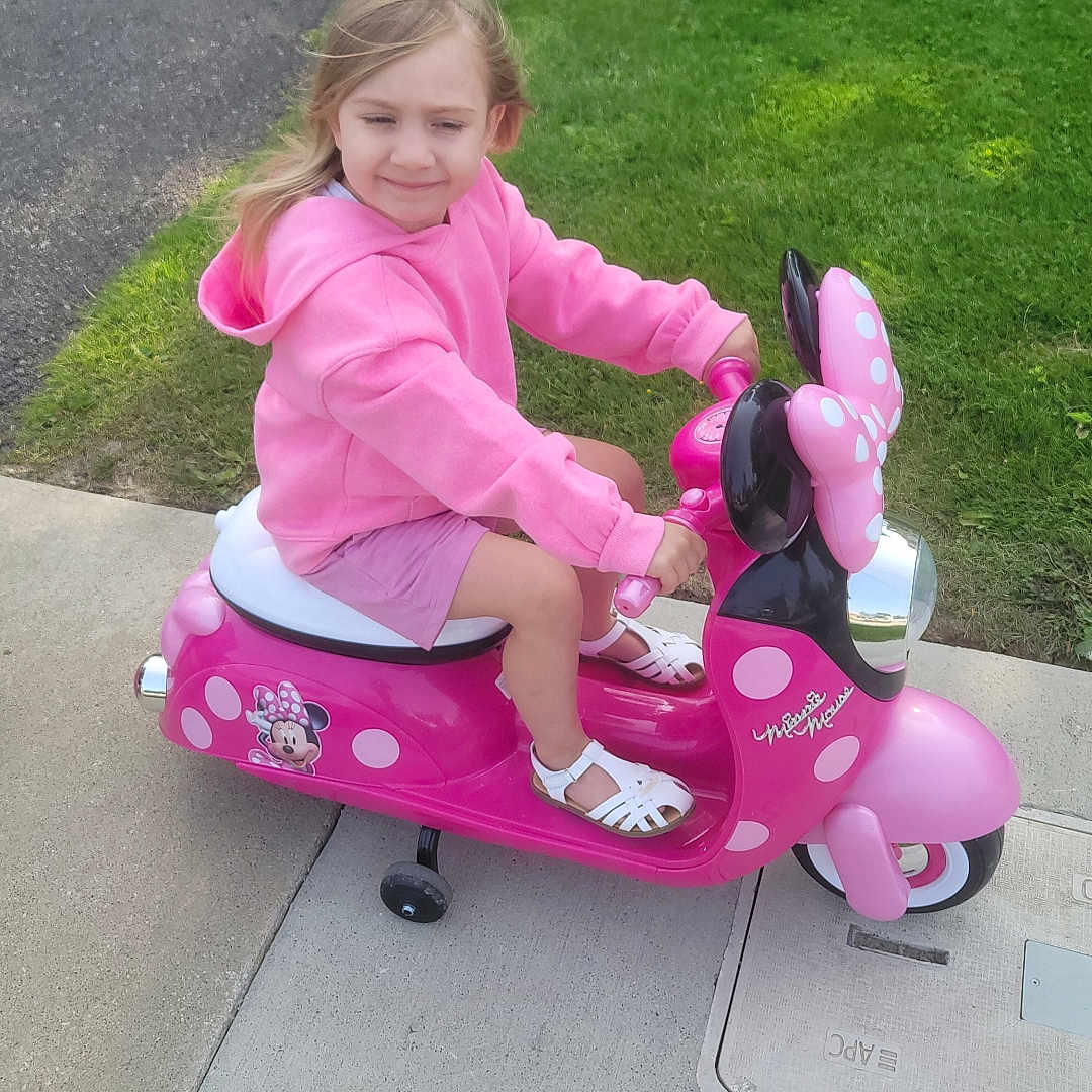 Destiny is registered to the contest to win money with this photo: casual_clothing, child, cute, daylight, fun, girl, grass, minnie_mouse, outdoor, person, pink_hoodie, play, recreation, shoes, sidewalk, smiling, toy_scooter, transportation, vehicle, white_sandals