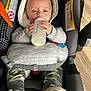 baby, bottle, camo_pants, car_seat, child, clothing, comfort, face, feeding, floor, hands, hooded_jacket, indoor, infant, milk, safety_labels, shoes, sitting, warm_clothes, wooden_floor