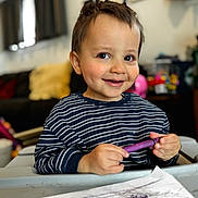 Eli is registered to the contest to win money with this photo: child, toddler, smiling, crayon, drawing, art, table, striped_shirt, indoor, living_room, cozy, face, happy, playful, person, scribbles, paper, toy, background, young